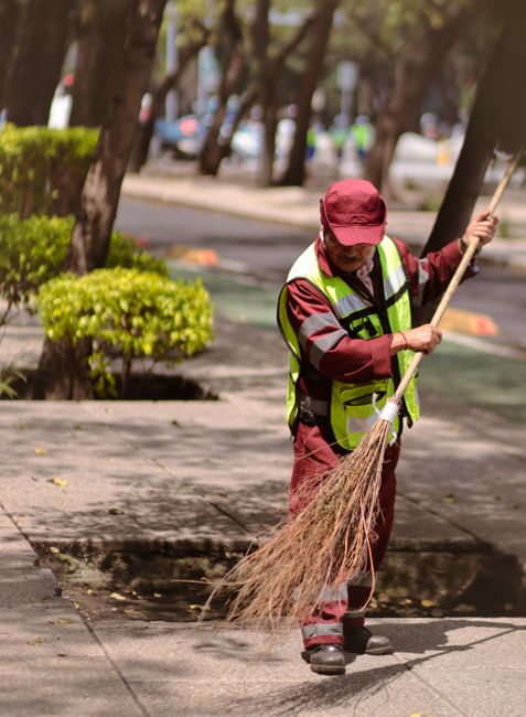 A street cleaner wearing a red cap, yellow high-visibility vest, and maroon uniform sweeping debris from a paved sidewalk with a traditional straw broom. The scene is set outdoors near a row of trees 